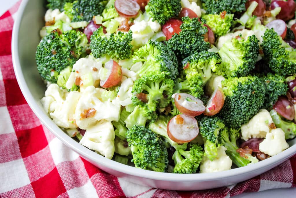 Broccoli, cauliflower salad with bacon and grapes in bowl with red and white checker towel.