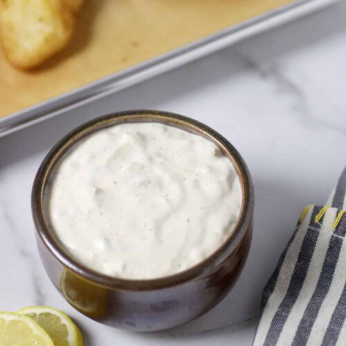 Tartar sauce in brown bowl with lemon, a pan of fried fish and striped napkin.