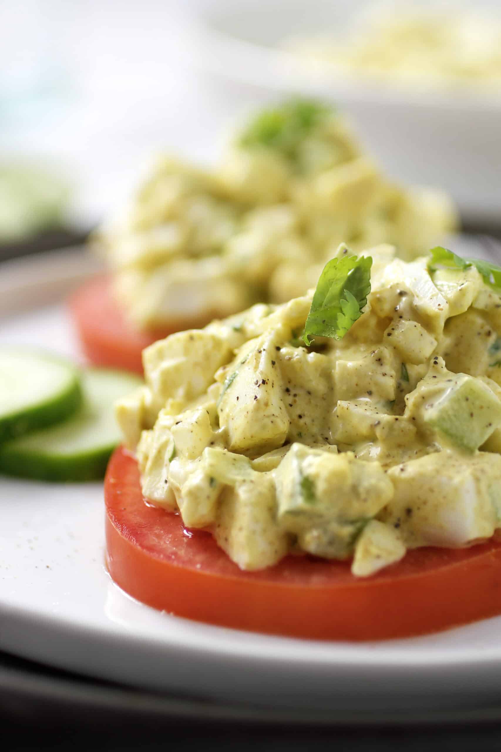 Egg salad on sliced tomato with cucumbers on white plate.