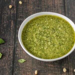 Basil parmesan pesto in bowl on table with pine nuts and fresh basil around bowl.