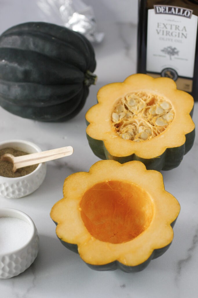 raw acorn squash cut in half with seeds removed in one of the two halves, salt and pepper on the side with a small measuring spoon, olive oil in the back and another whole acorn squash