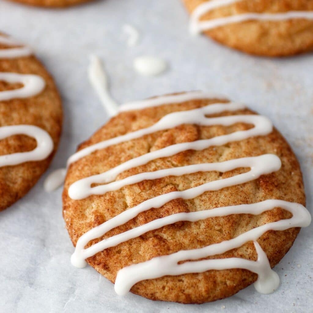 Cinnamon sugar coated cookie with icing drizzled over the top on parchment paper with other cookies.