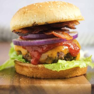 cheese burger with a grilled bun, bacon, lettuce, onion and ketchup on a wooden cutting board