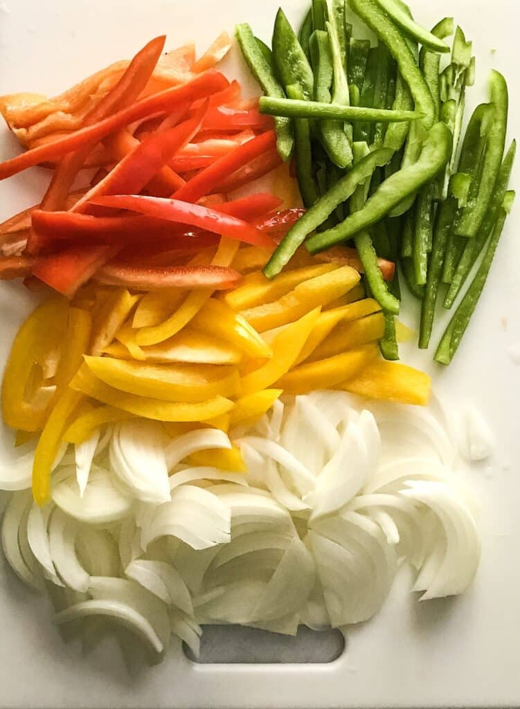 Sliced ed, green and yellow peppers and sliced white onion on cutting board.