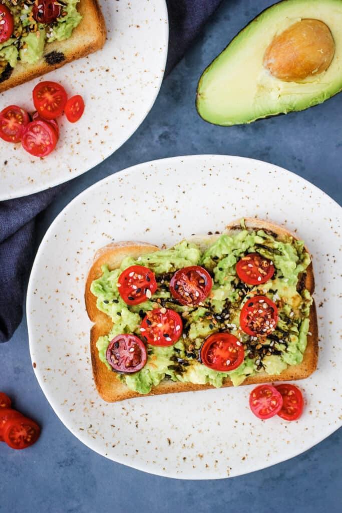 Avocado Toast on plate with extra tomatoes and half an avocado above the plate.