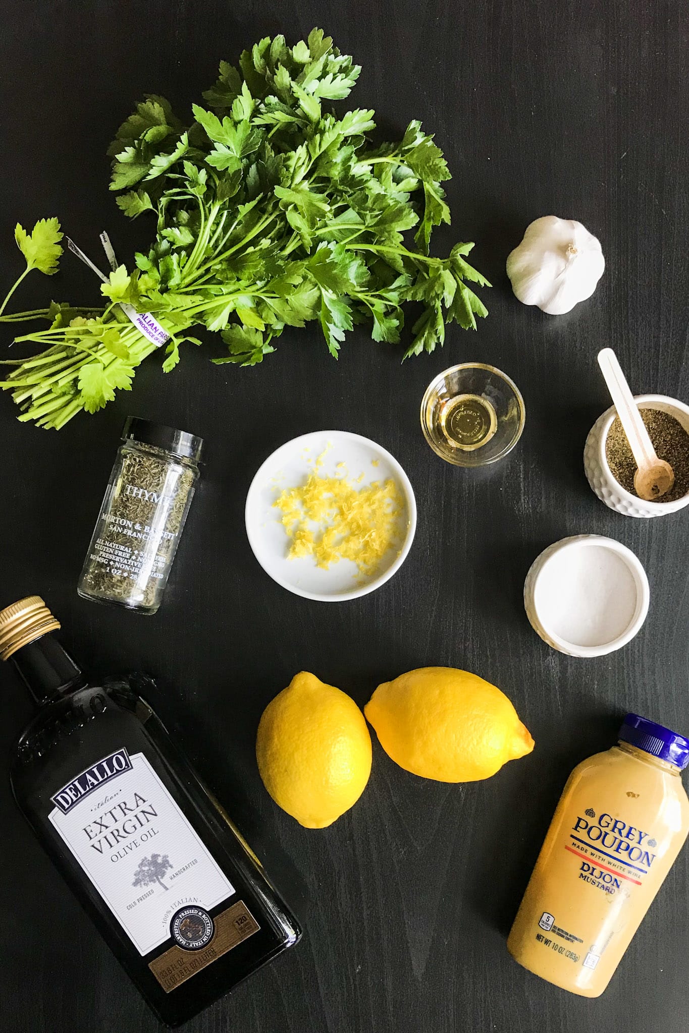 All the ingredients needed to make simple lemon vinaigrette recipe.  Two lemons, dijon mustard, olive oil, salt, fresh parsley, thyme, pepper, lemon juice and zest, garlic and honey.