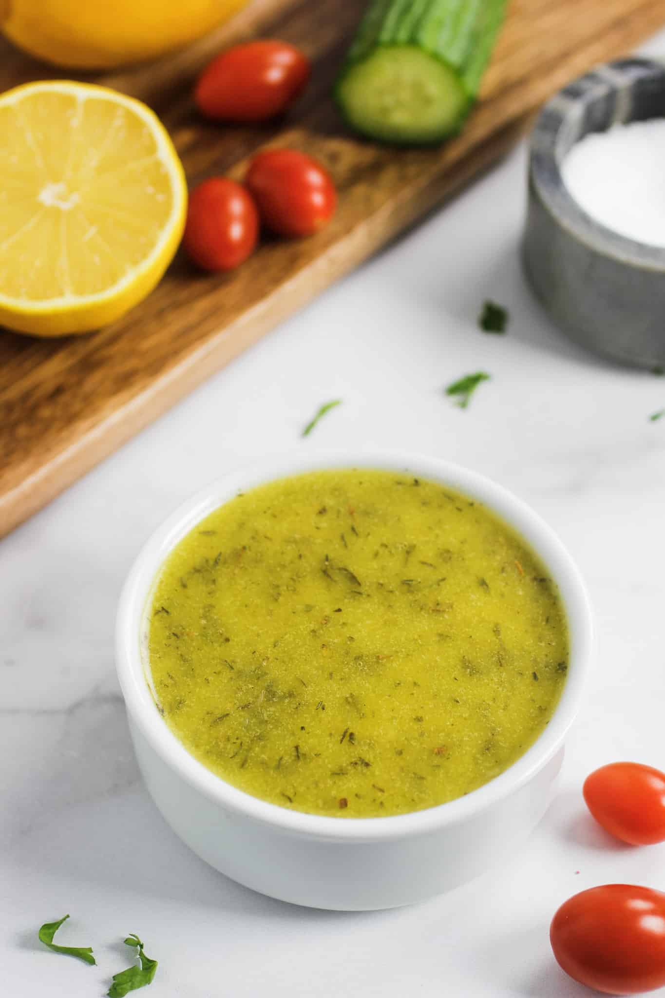 Simple Lemon Vinaigrette in a white bowl with vegetables on a cutting board and cherry tomatoes on the table.