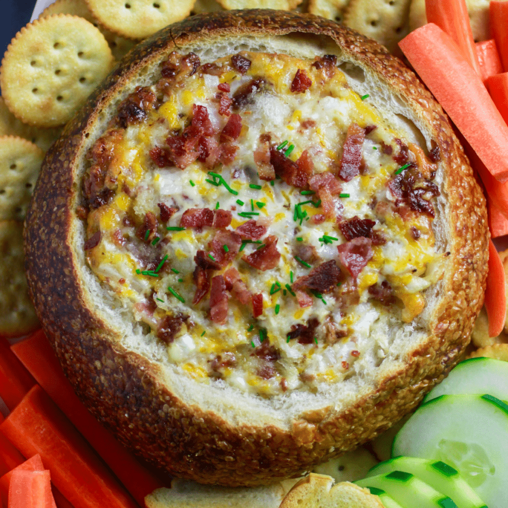 cheesy bacon dip recipe in a bread bowl with crackrs and vegetables.