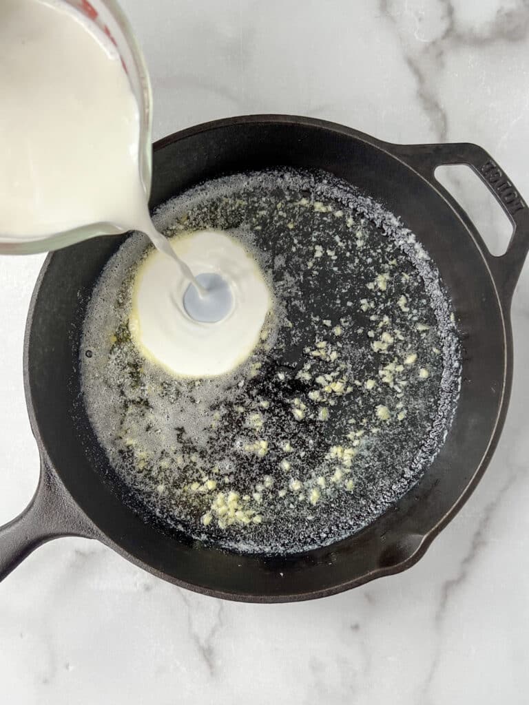 cast iron skillet with melted butter and minced garlic with heavy whipping cream being poured into pan.