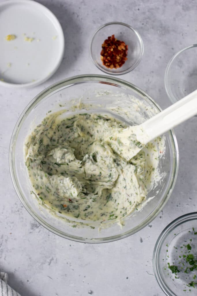 Butter with herbs in a clear mixing bowl with a white spatula.