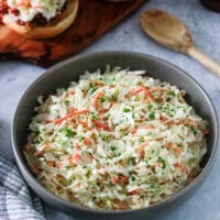 Sweet Creamy Coleslaw in a gray bowl with two bbq sandwiches and a wooden spoon.