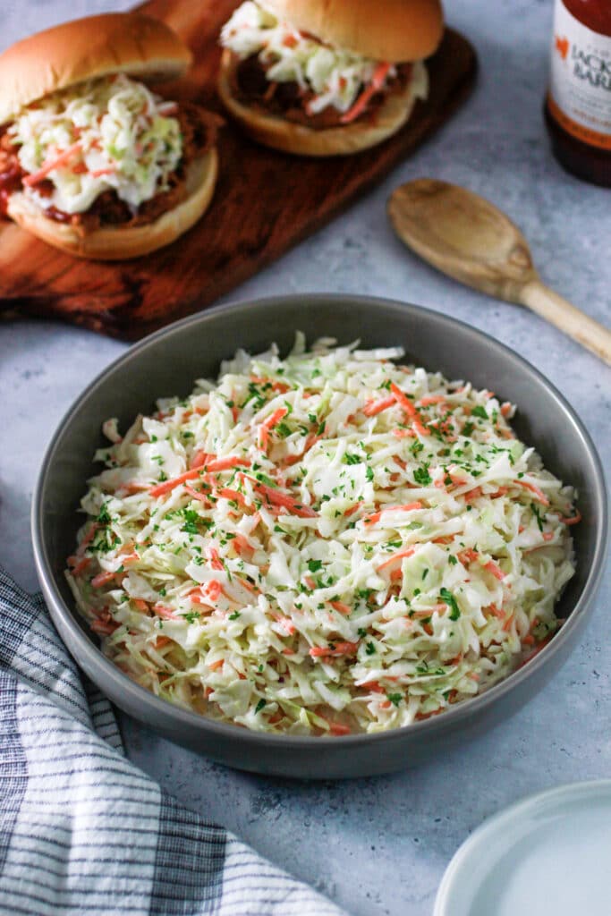 Sweet Creamy Coleslaw in a gray bowl with two bbq sandwiches and a wooden spoon.