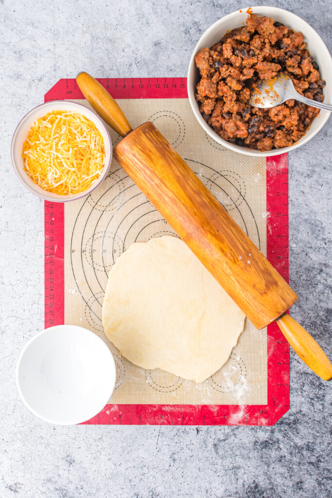 Rolling pin with pie dough, ground beef mixture and cheese on a cutting board.
