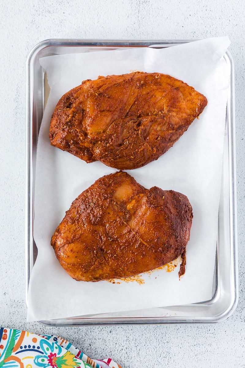 Chicken breasts that have been marinated on a baking sheet with parchment paper before baking.