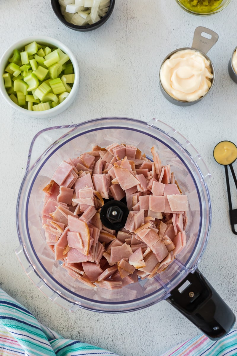 Roughly chopped ham in food processor with bowls of other ingredients around the processor bowl.