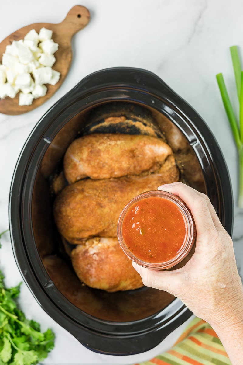 Salsa being poured over seasoned chicken breasts in a slow cooker.