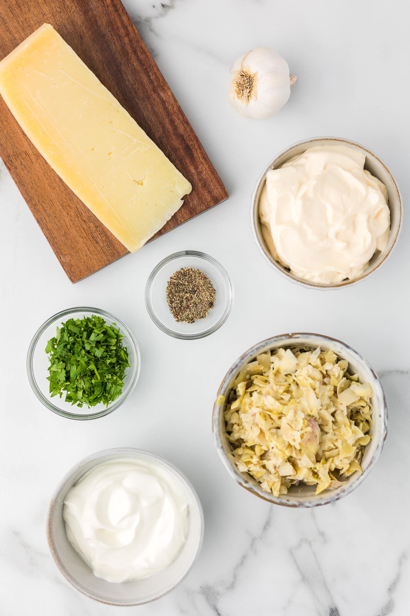 Ingredients to make hot artichoke dip including, garlic, parmesan cheese, mayonnaise, black pepper, parsley, sour cream and artichoke hearts.