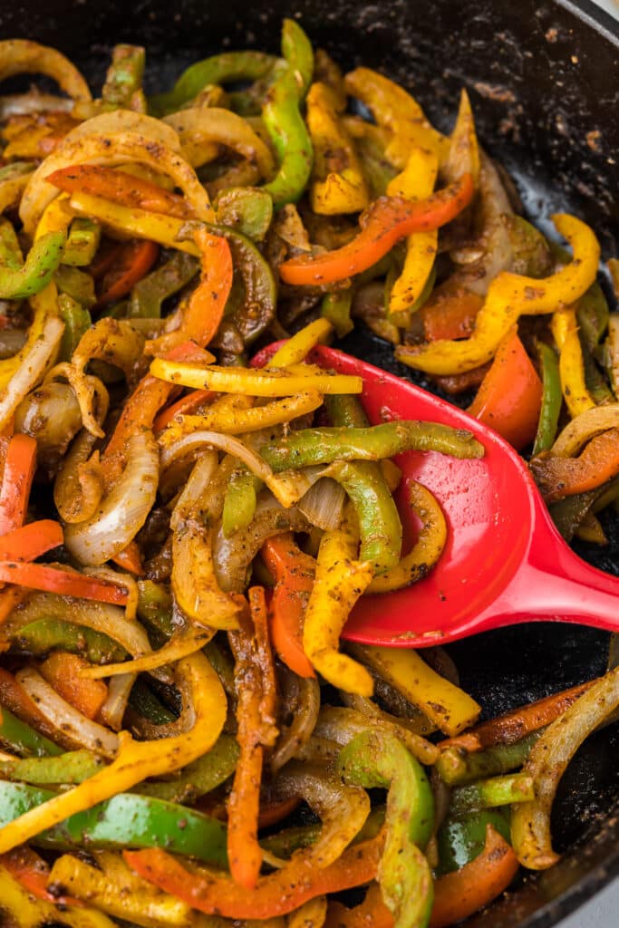 Fajita style onion and peppers with a red spoon in a black skillet.
