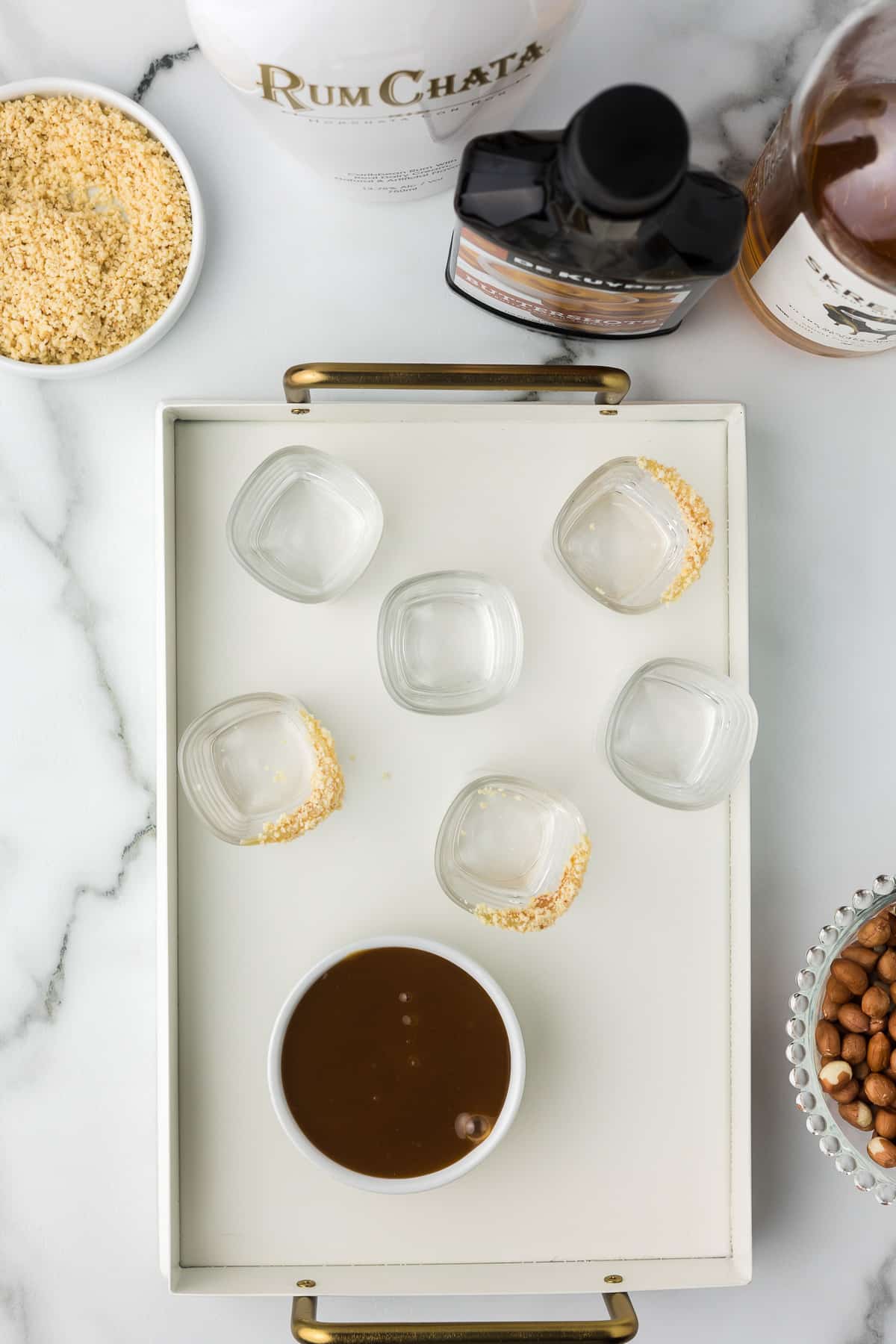 A tray with shot glasses prepared with caramel and chopped peanuts and a bowl of caramel sauce.
