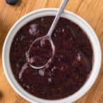 Blueberry puree in a white bowl with a spoon on a cutting board.