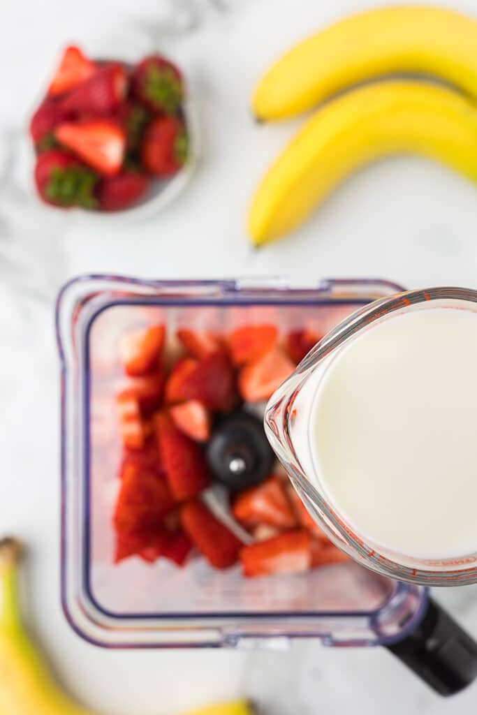 Strawberries and frozen bananas in a blender with milk being added.