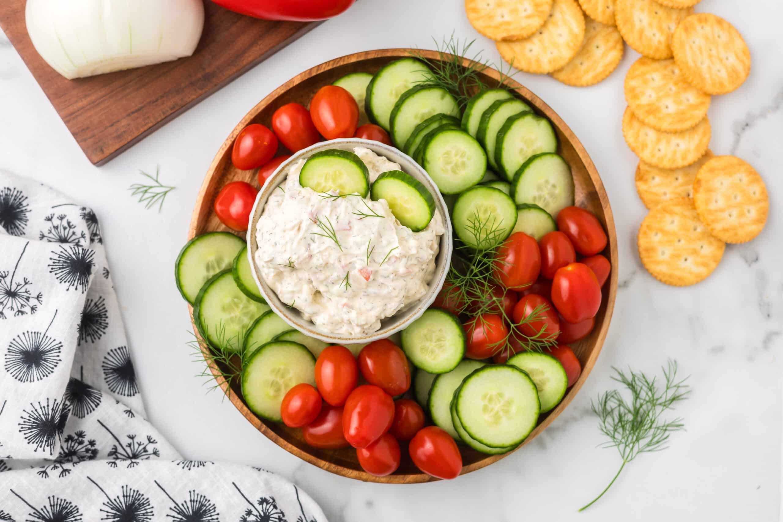 A platter with fresh cucumber slices and cherry tomatoes surrounding a bowl of creamy dip, garnished with dill. Crackers and a kitchen towel with a floral pattern are nearby on a marble surface.