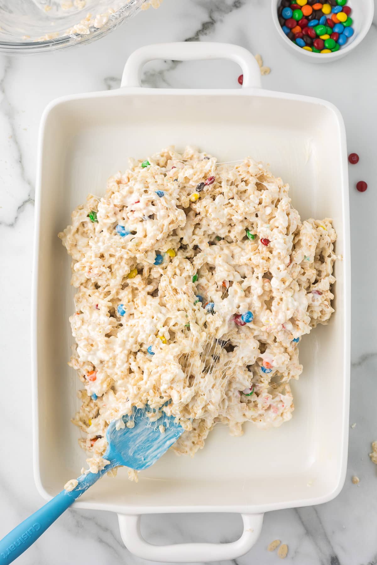 9 X 13 white pan with M&M rice krispie treats being poured in with a blue spatula.