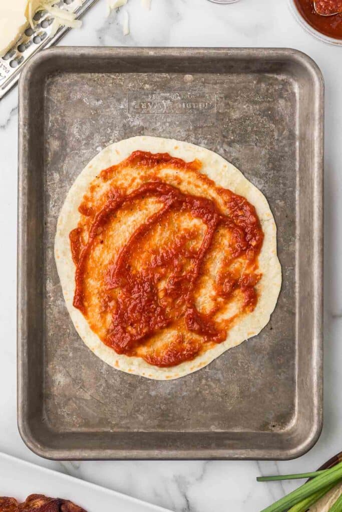 A tortilla on a baking sheet with pizza sauce.