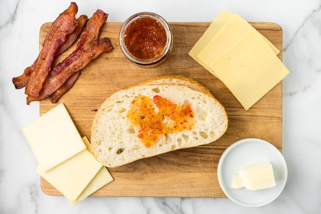 Bread with bacon, jelly and cheese on a cutting board.