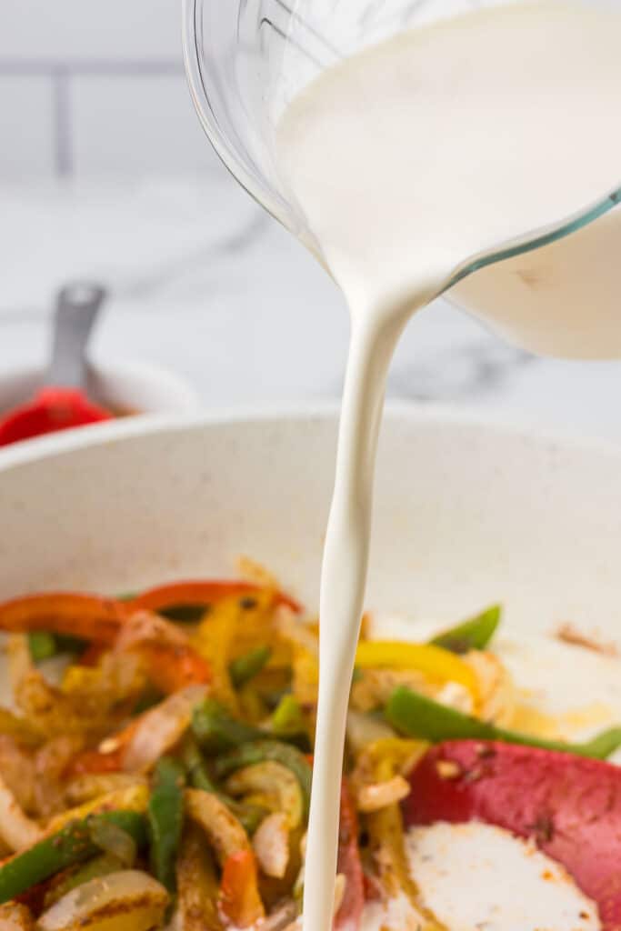 Heavy whipping cream being poured into a pan of onions and bell peppers.