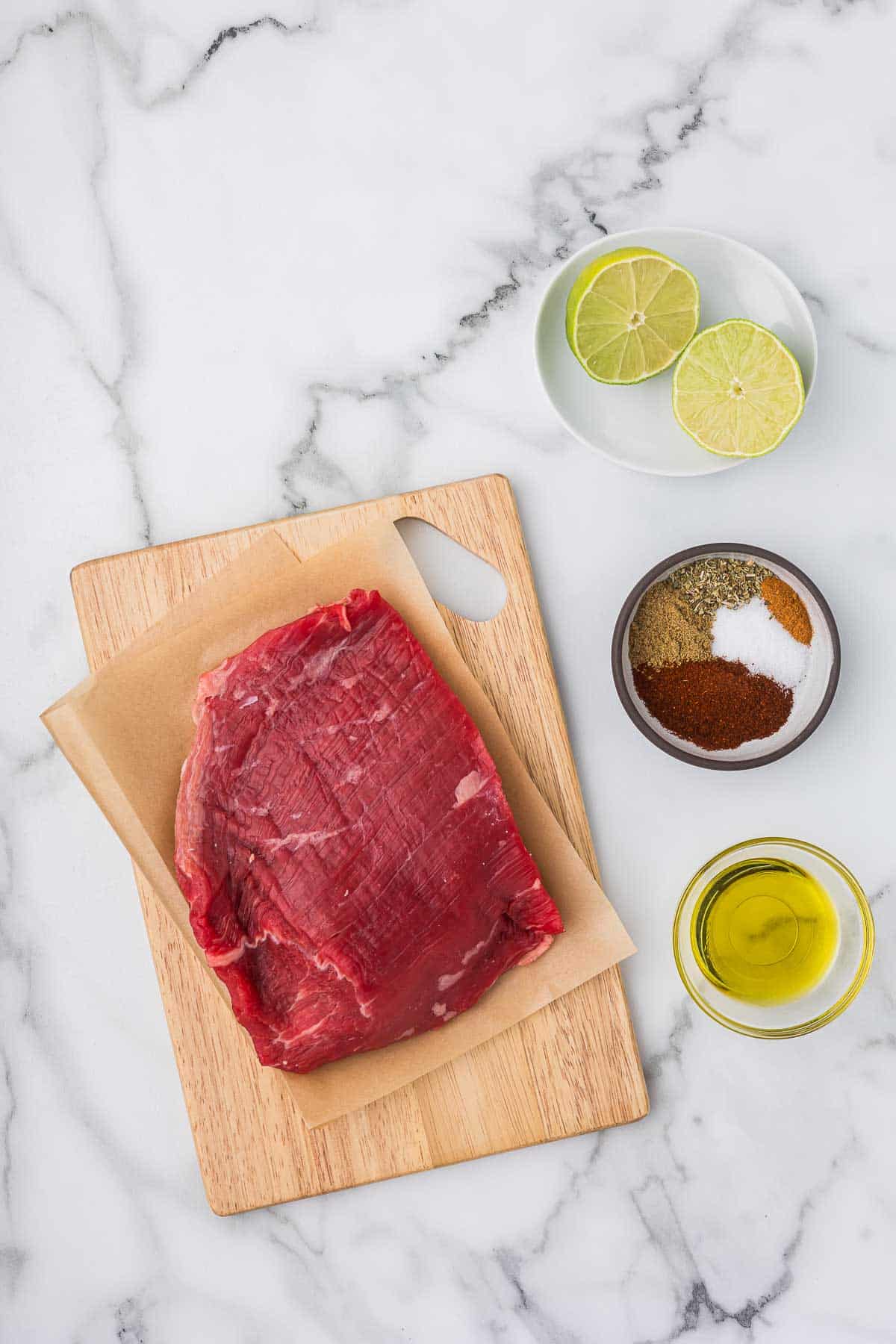 Raw flank steak on a wooden cutting board, olive oil, spices, and limes. 