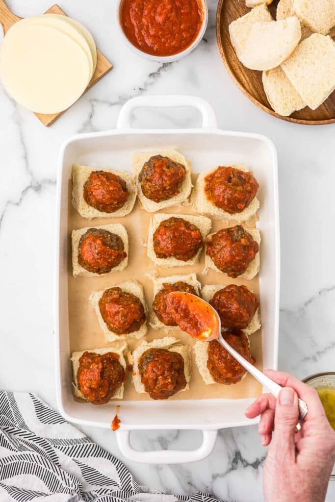 A white baking dish with  the bottom roll, a meatball and marinara sauce being added to the top of the meatball.
