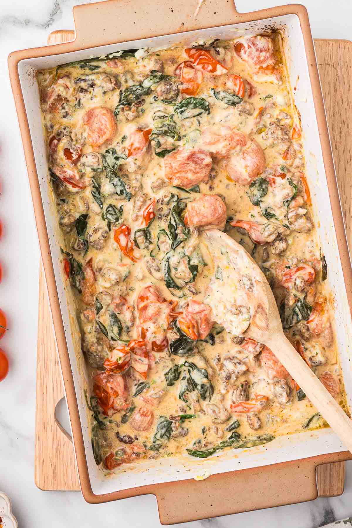 Baked Boursin cheese dip with cherry tomatoes and fresh basil with a wooden mixing spoon on a wooden cutting board.