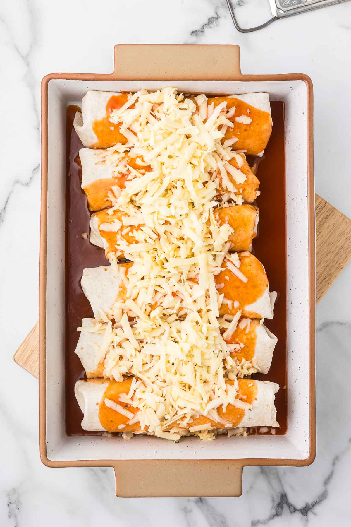 Pulled Pork burritos, enchilada sauce, hot sauce and shredded cheese in a casserole dish on a wooden cutting board.