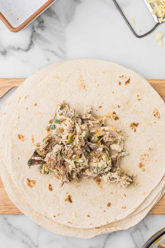 Pork mixture in the middle of a flour tortilla on a wooden cutting board, cheese on a cheese grater. 