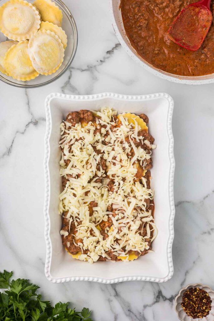 A casserole dish with ravioli, sauce and cheese before the second layer of ingredients is added.