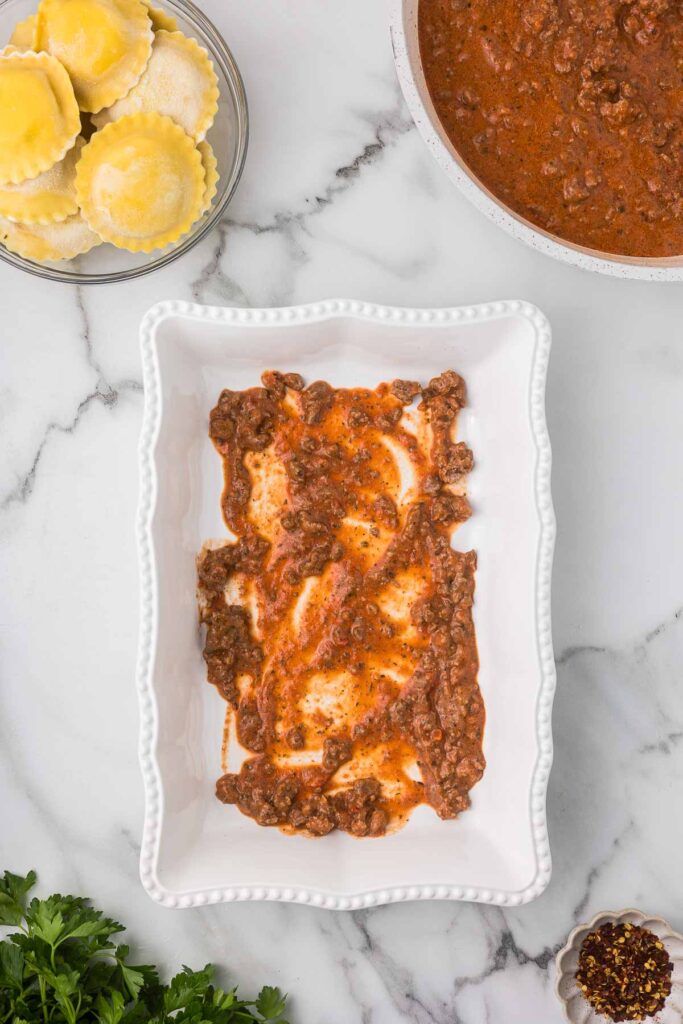 Casserole dish with 1/4 cup of meat sauce before a single layer of ravioli is added.