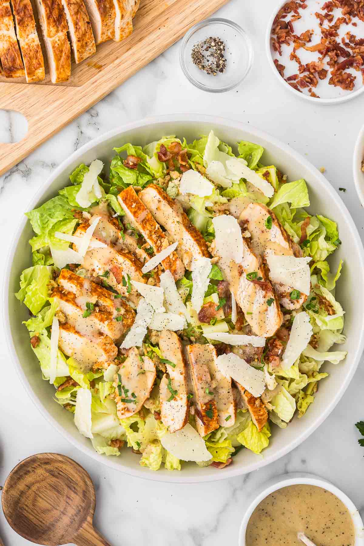 Caesar salad in a white bowl with bacon, shaved parmesan cheese and sliced chicken breast.