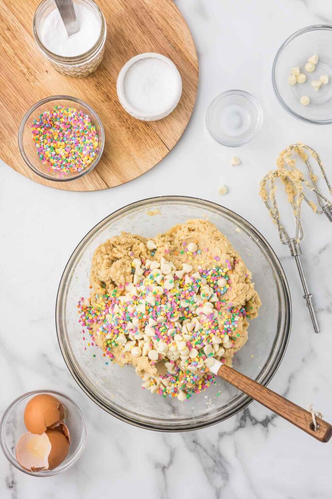 Sprinkles, butter, white sugar, brown sugar, baking powder, flour, milk, eggs, salt, brown sugar and white chocolate chips in a bowl with blender whisk attachments. 