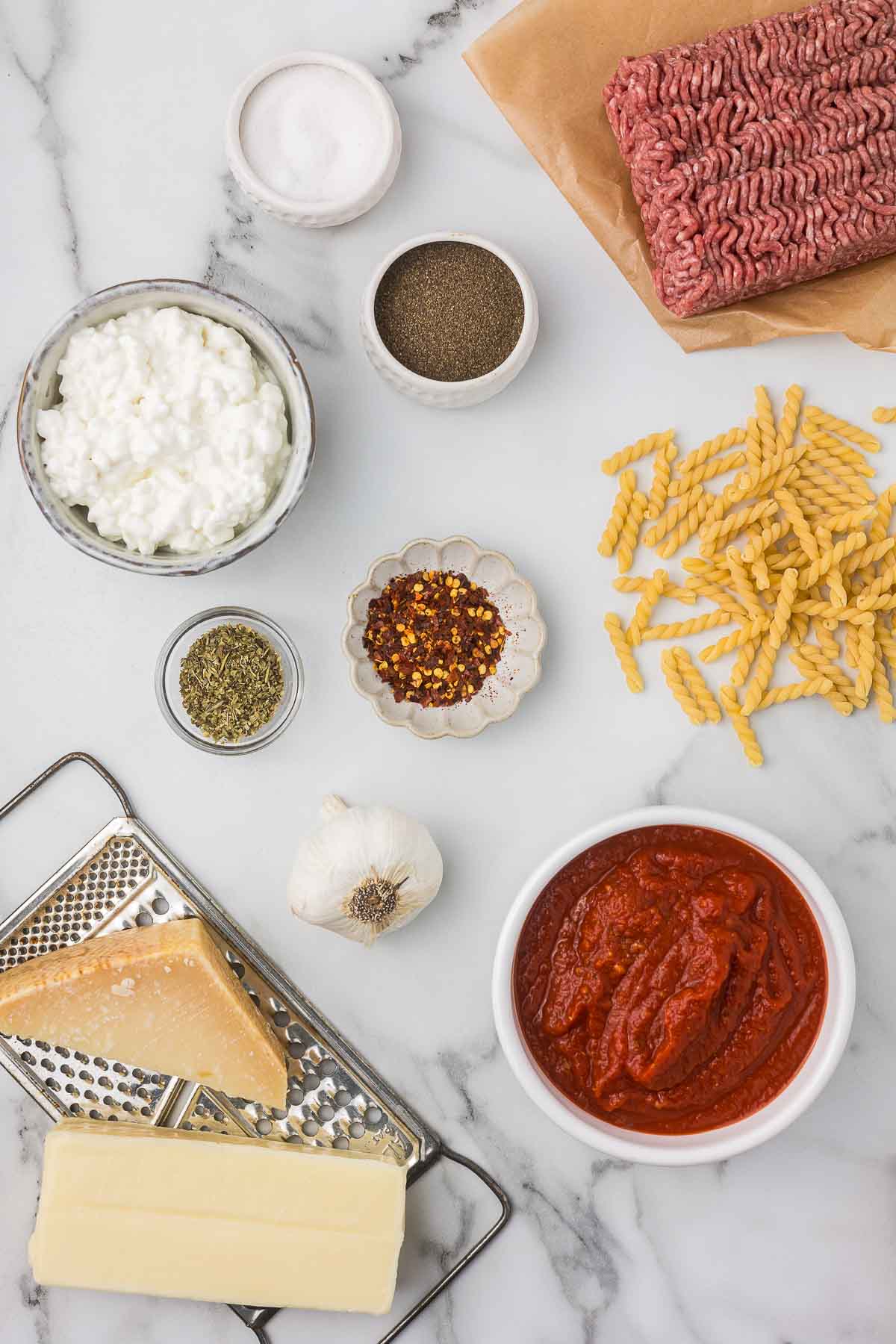 Ground beef, salt, pepper, chili flakes, Italian seasoning, cottage cheese, spiral pasta, garlic, marinara sauce, mozzarella cheese and parmesan cheese on a cheese grater. 