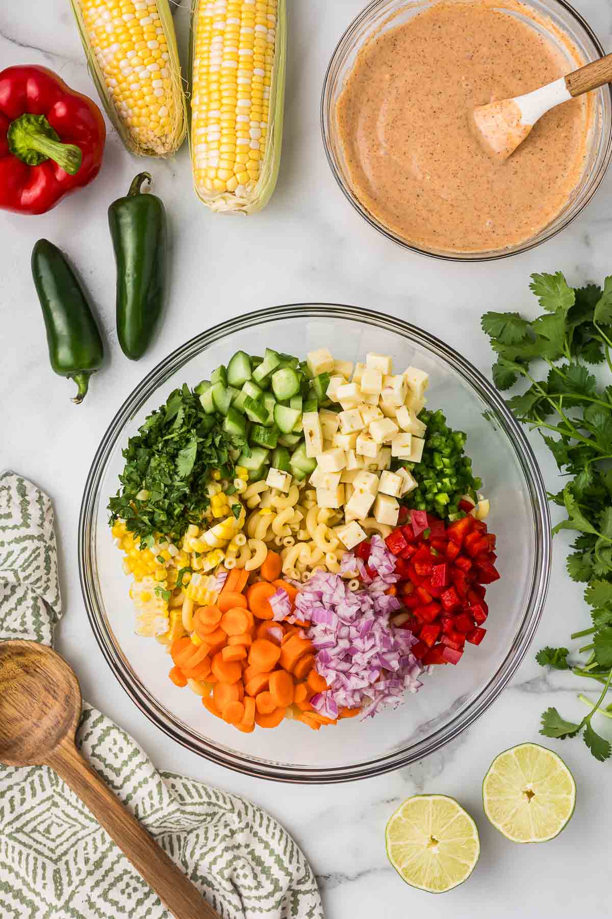 A glass bowl with pasta, onion, bell pepper, cucumbers, carrots, cilantro and jalapenos.