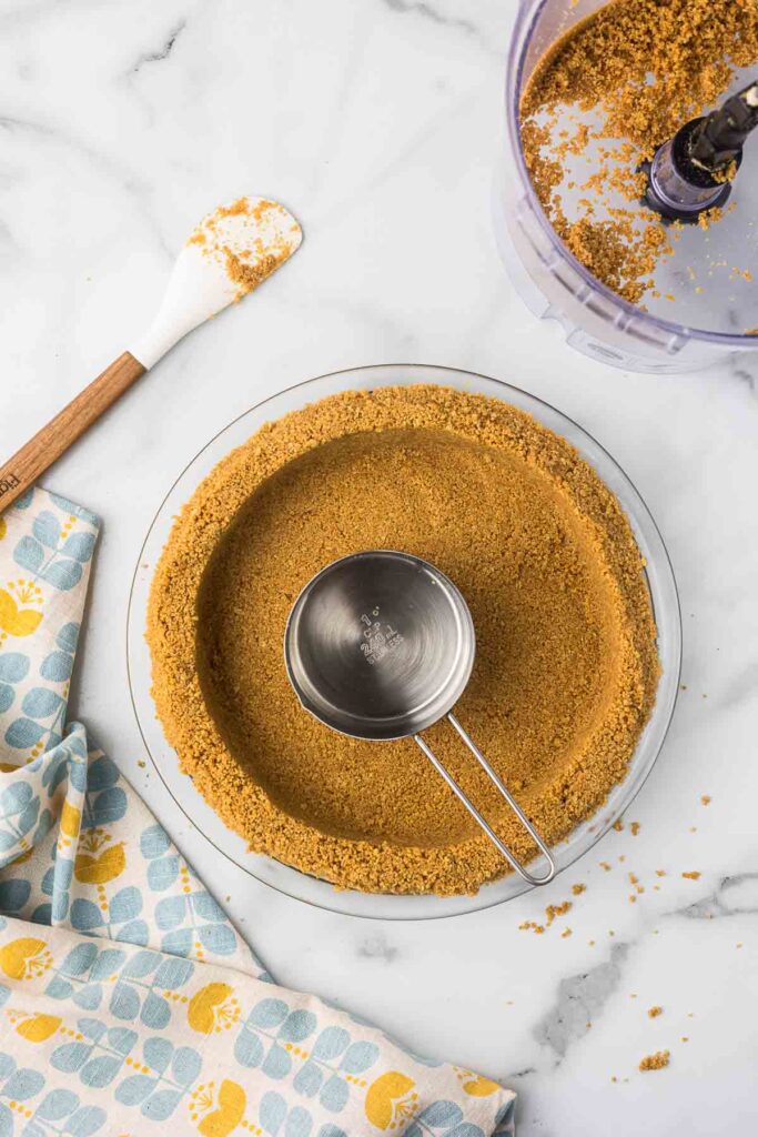 Graham cracker crust in a glass pie plate being pressed down with a measuring cup.