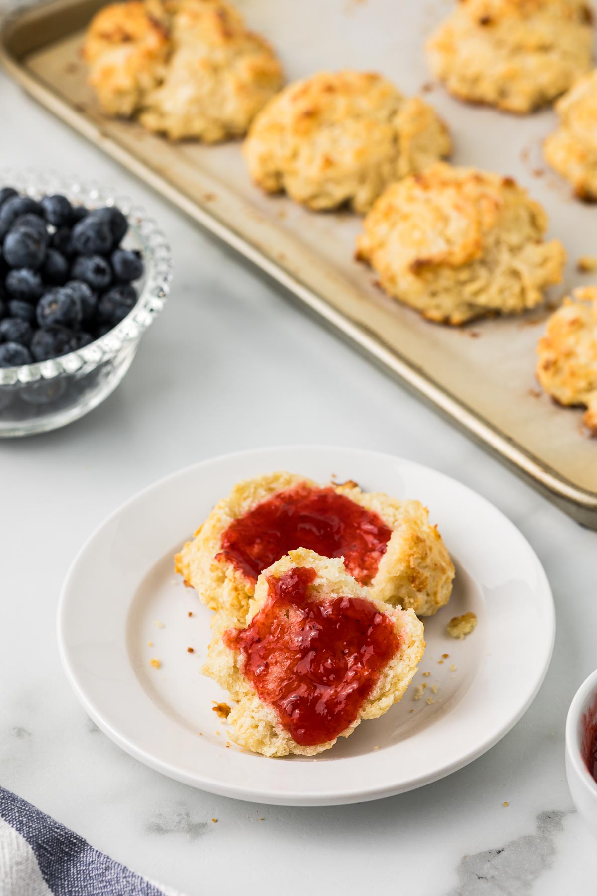 Buttermilk biscuits on a parchment paper lined cookie sheet, bowl or blueberries, a biscuit with strawberry jam