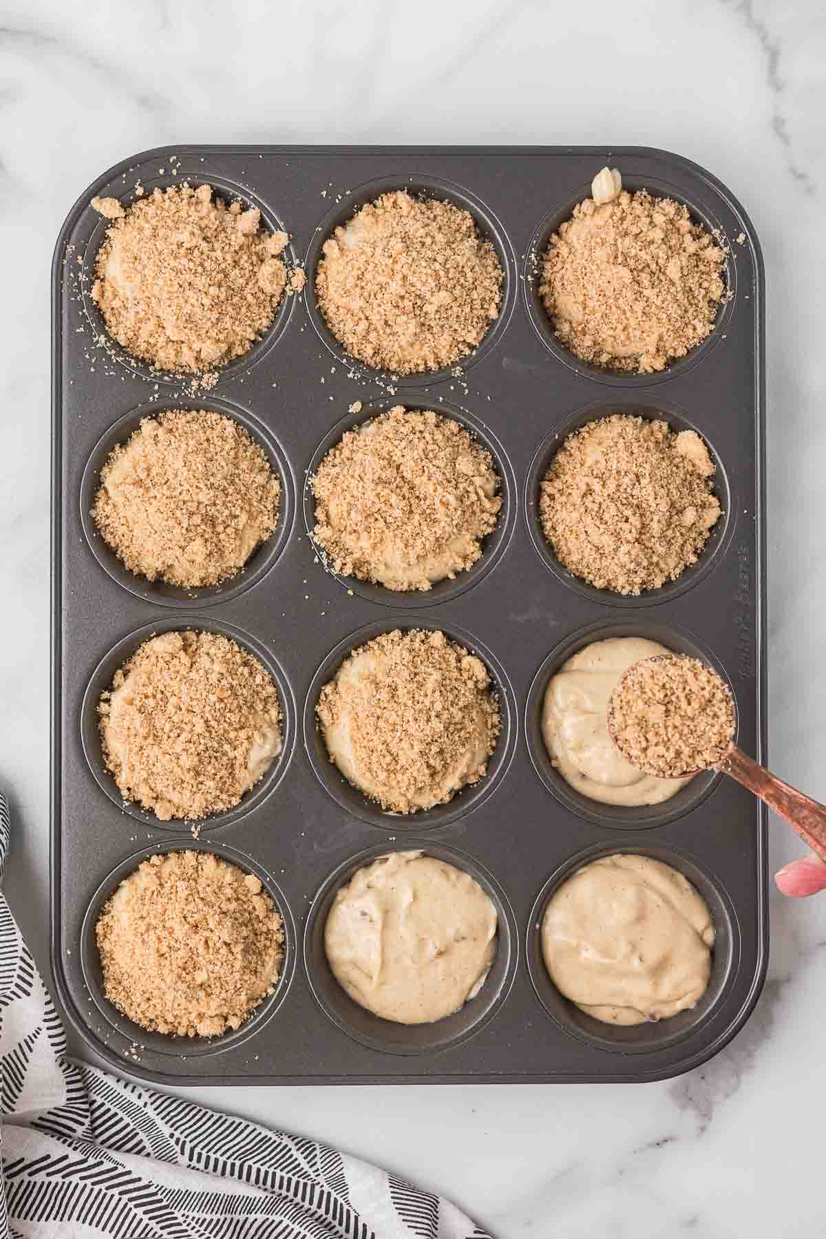Muffin batter in a muffin pan with the crumb topping being added.