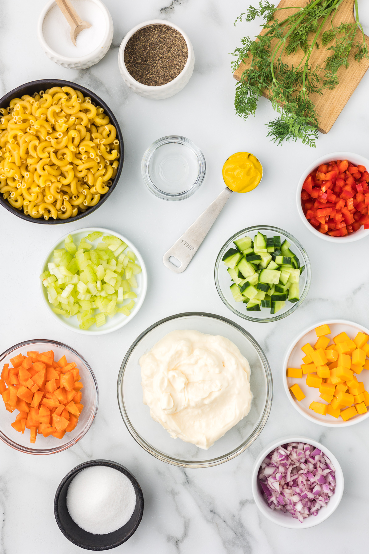 Individual bowls with sugar, red onion, carrots, cubed cheddar cheese, diced celery and cucumber, mayonnaise, salt and pepper, macaroni, fresh dill, vinegar and yellow mustard.