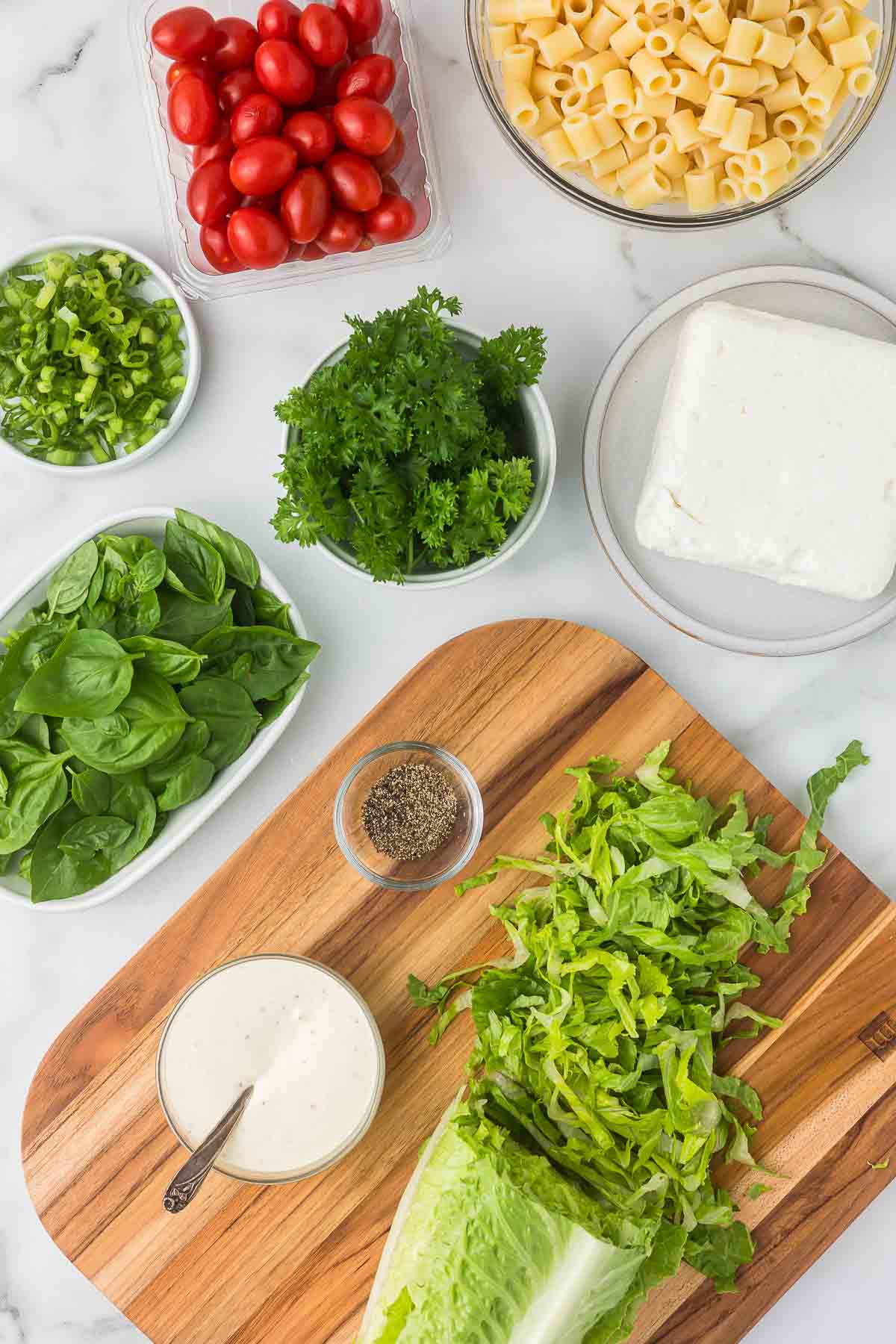 Ingredients to make caesar pasta salad including feta cheese, pasta, fresh basil and parsley, green onions lettuce and Caesar dressing.