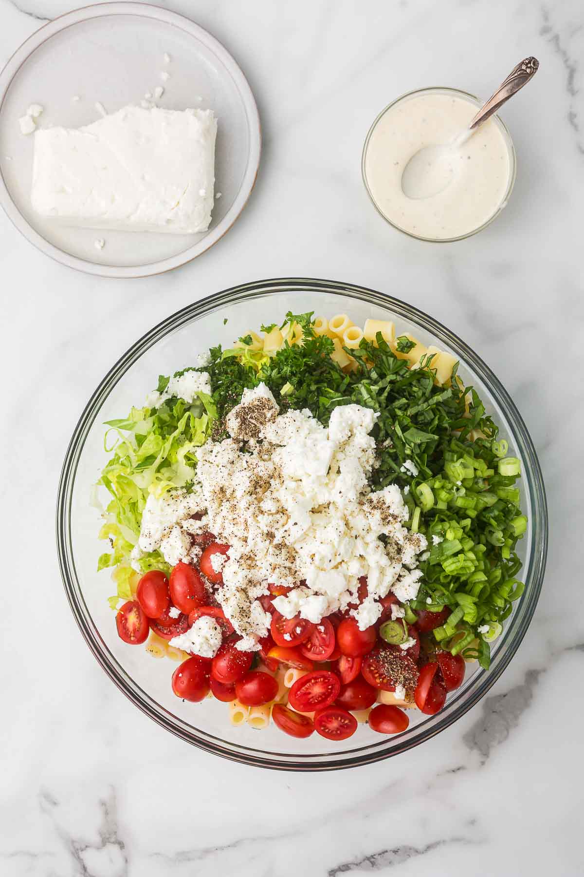 Pasta, fresh basil, parsley, romaine lettuce, tomatoes and feta cheese in a bowl before adding Caesar dressing to make pasta caesar salad.