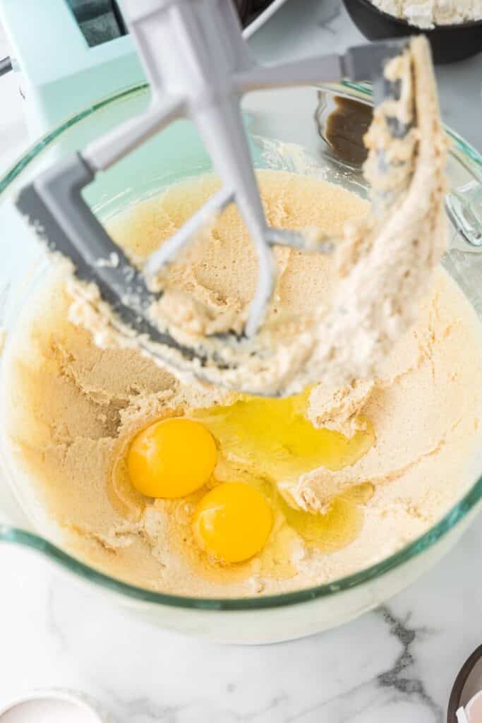 Eggs being added to a glass kitchen aid bowl with ingredients to make oreo choclate chip cookies.