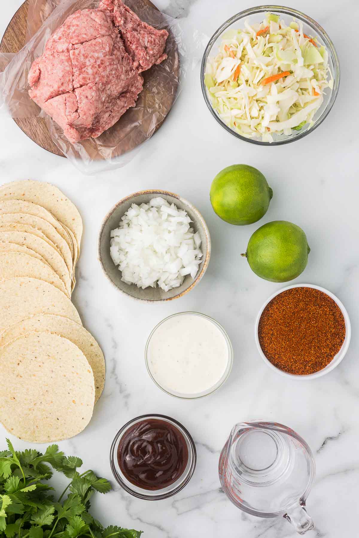 Ingredients to make bbq tacos including shells, onion, lime, cilantro, bbq sauce, water, ranch dressing, ground pork and slaw.