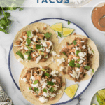 Top view of a plate with three BBQ pork tacos, garnished with cheese and cilantro. Lime wedges are on the side. The plate sits on a patterned white surface, with chopped onions and sauce nearby. Text above reads "MUST TRY BBQ PORK TACOS.